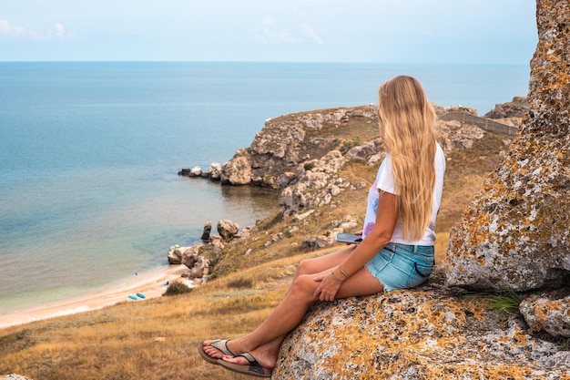 A woman with long blond hair sits on top of a mountain and looks down at the sea below travel and