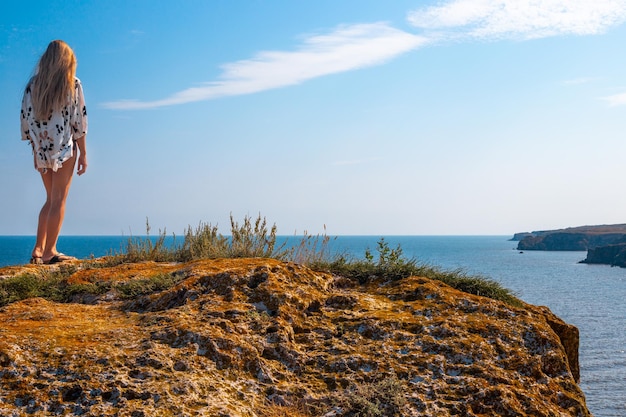 Женщина с длинными светлыми волосами стоит на краю горы и смотрит на морской пейзаж Путешествия и туризм