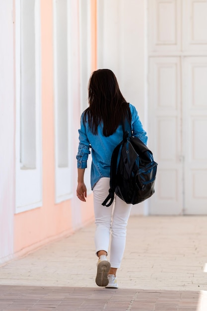 Woman with long brown hair walking with backpack View from the back White jeans and blue shirt Light hall background