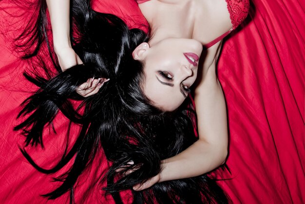 Woman with long brunette hair and fashionable makeup on face on red bed in bedroom