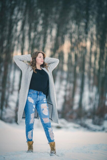 Woman with long brunette hair wearing jeans with a cardigan in a forest covered in the snow