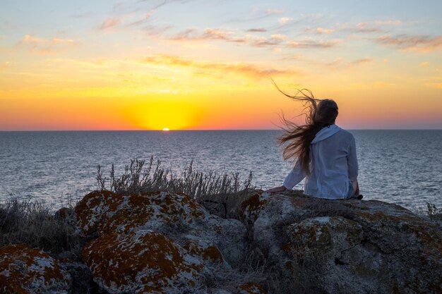 Женщина с длинными волосами сидит на камне на вершине горы на морском побережье при заходе солнца задний вид Путешествия и туризм