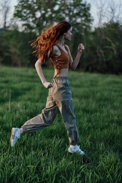 A woman with long red hair works out and runs on the green grass in the park in sweatpants and sneakers