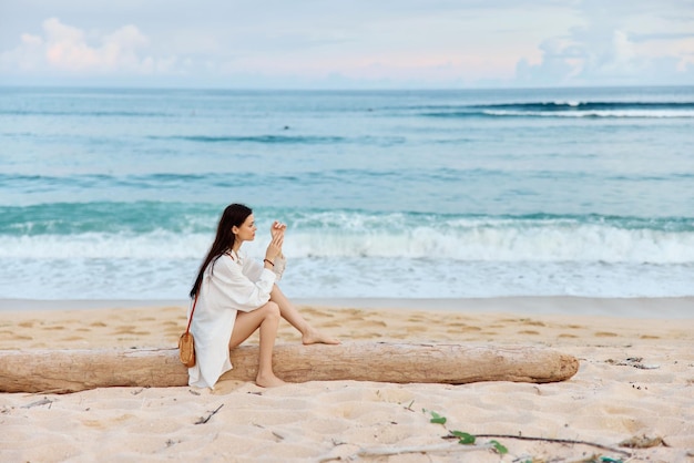 Женщина с длинными мокрыми волосами после купания сидит на песке спиной к камере и смотрит на закат у океана на острове Бали