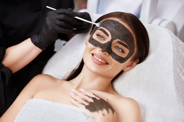 Женщина с прекрасной улыбкой, получающая уход за лицом с угольной маской косметологом в спа, клиентка салона красоты, имеющая процедуры для лица
