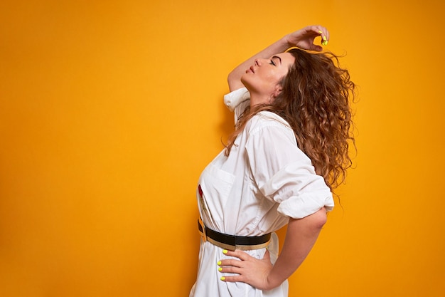 A woman with lush curly hair stands sideways. She holds his hair back with one hand, straightens it, and looks away.