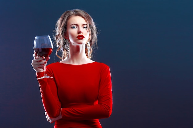 Woman with makeup, hairstyle wearing red dress posing with glass of vine over dark background