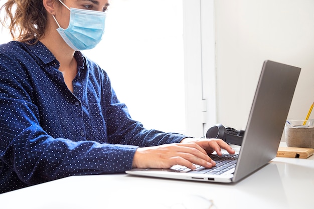 Женщина с маской, одетая в синее, работает со своим ноутбуком на белом столе в ярком офисе