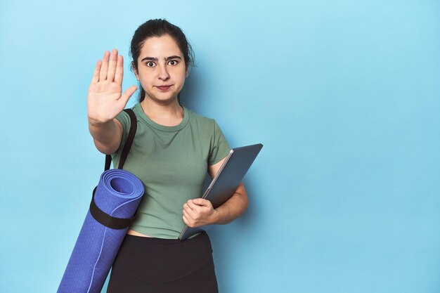 Woman with mat and laptop on blue standing with outstretched hand showing stop sign preventing you