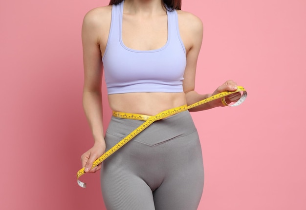 ピンクの背景のクローズアップで彼女のスリムな体を示す測定テープを持つ女性