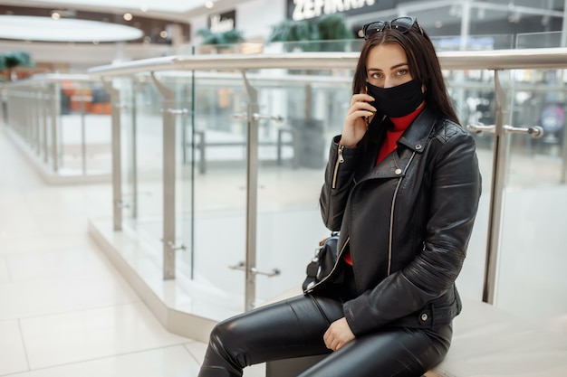 Woman with medical black mask and mobile phone in a shopping\
center. coronavirus pandemic. a woman with a mask is standing in a\
shopping center. a woman in a protective mask is shopping at the\
mall