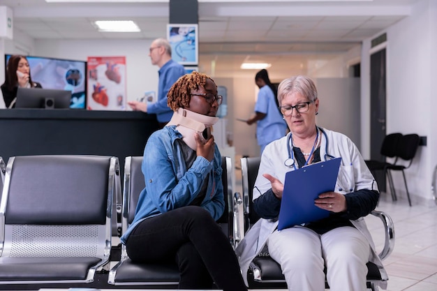 Woman with medical neck collar at examination with doctor,\
sitting in waiting room lobby. patient with cervical foam and\
senior medic filling in checkup report papers at appointment in\
hospital.