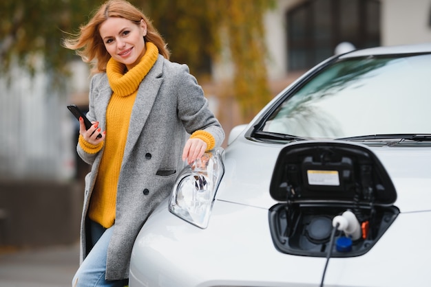 電気自動車の充電の近くで携帯電話を持っている女性。屋外の公共充電ステーションでの車両充電。カーシェアリングのコンセプト