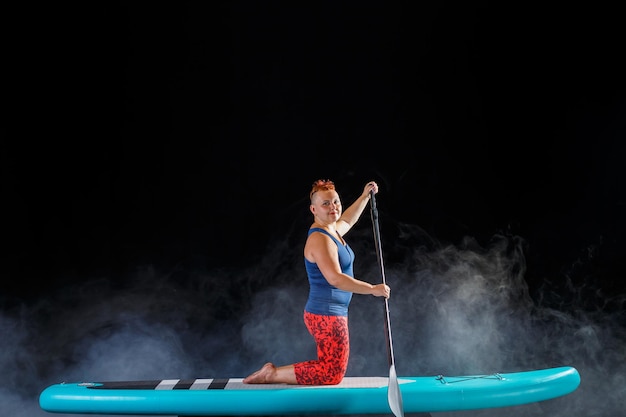 A woman with a mohawk on a sup board on her knees with an oar in the fog on a black background