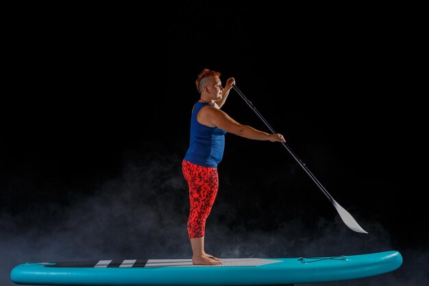 A woman with a mohawk on a sup board kneeling with an oar on a black background