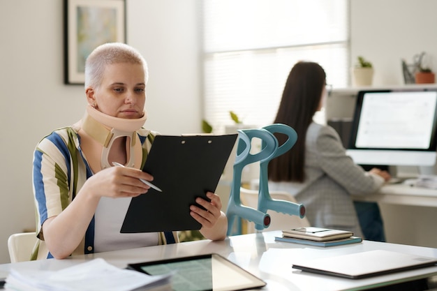 Woman with neck brace sitting at table in office of social service and examining contract