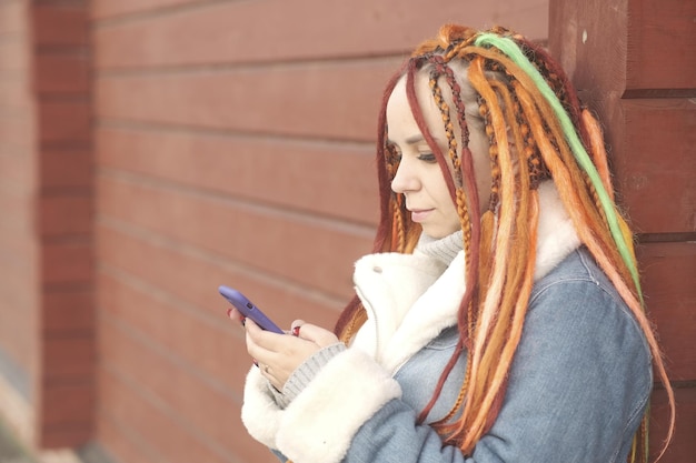 オレンジ色のドレッドヘアを持つ女性と赤い木製の背景にスマート フォンを使用して暖かいデニム ジャケットの女性