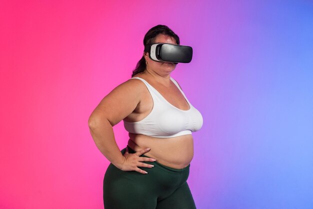 青と赤の背景のスタジオでポーズをとるトラック スーツと VR ヘッドセットを着た太りすぎの女性