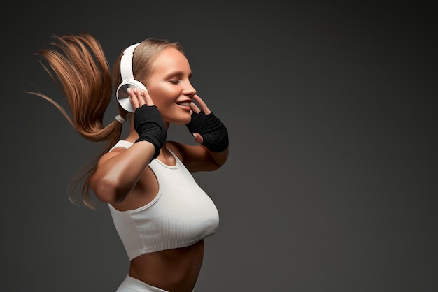 Woman with perfect abs wearing headphones and white clothes standing in gym isolated grey background