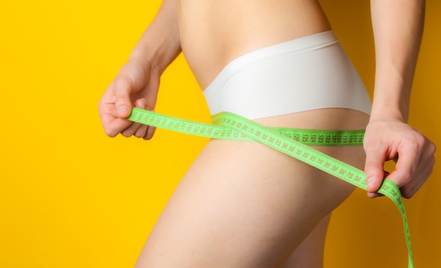 A woman with a perfect body in panties is measuring her hips with ruler on yellow