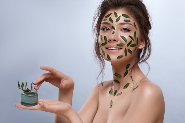 Woman with perfect skin having green leaves on her face and holding cream