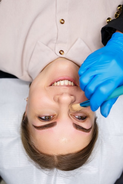 Woman with permanent makeup tattoo on her eyebrows. Close-up beautician makes makeup applies a foundation. Professional make-up and cosmetic skin care.