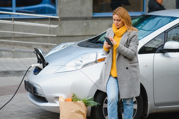 レンタル電気自動車の近くに電話を持つ女性。充電ステーションで充電された車両。