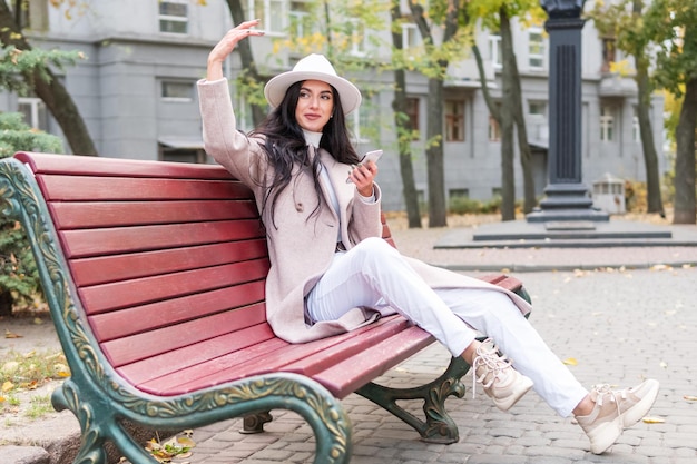 woman with a phone sits on a bench and waves her hand beckons to her