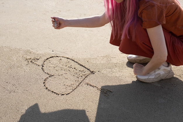 woman with pink hair draws a heart with a stick on the sand