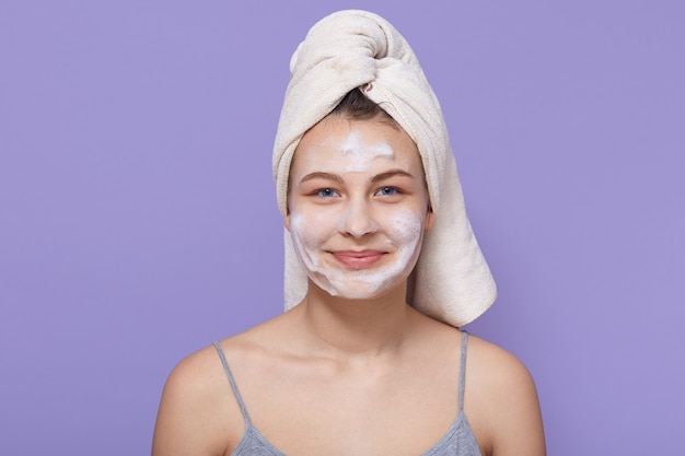 Woman with pleasant appearance, being wrapped in white towel and white facial mask on face