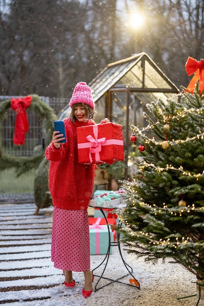 屋外のクリスマスツリーの近くにプレゼントと電話を持つ女性