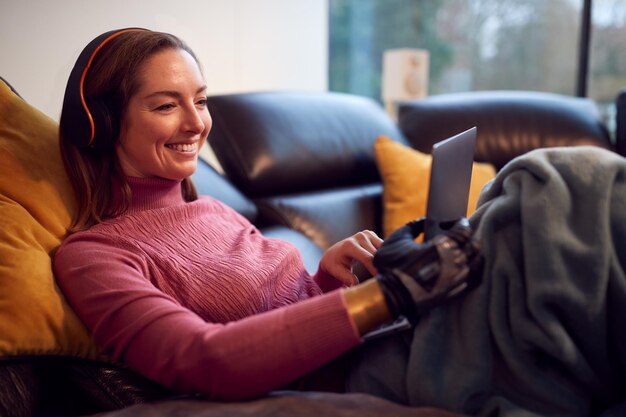 Photo woman with prosthetic arm wearing wireless headphones and watching movie on laptop on sofa at home