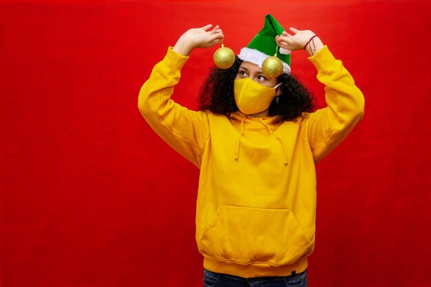 Woman with protective mask and Santa hat has spheres of Christmas tree decoration in her hands