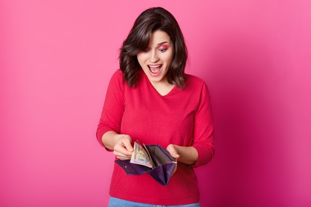 woman with purple wallet full of money in hands looks astonished. Surprised woman wears shirt, holds opened purse