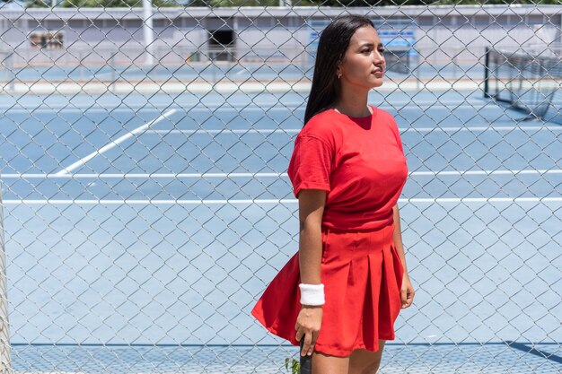 Woman with racket and ball standing near tennis court fence during break in training