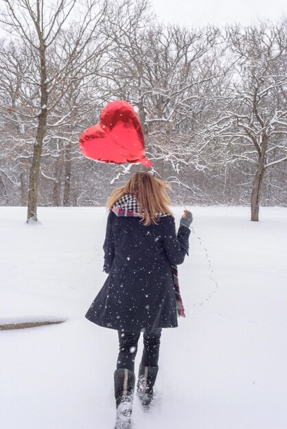 Woman with red balloon walking on snow covered road against bare trees