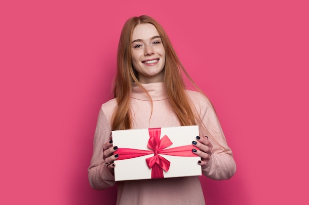 woman with red hair and freckles is showing at camera present box well packed posing and smile on a red wall