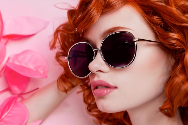 A woman with red hair wearing sunglasses and a pink background