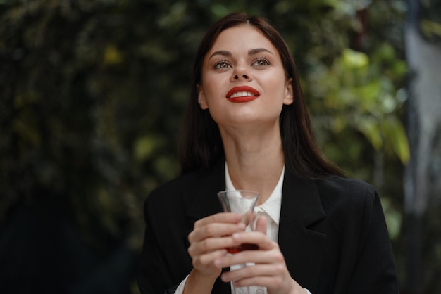 Женщина с красными губами улыбается с зубами пьет чай в кафе из турецкой стеклянной кружки в белой рубашке и черной модной куртке на улице летние путешествия каникулы в городе