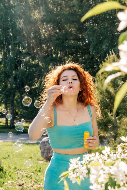 A woman with red lush hair blows soap bubbles in the spring during the flowering of apple trees