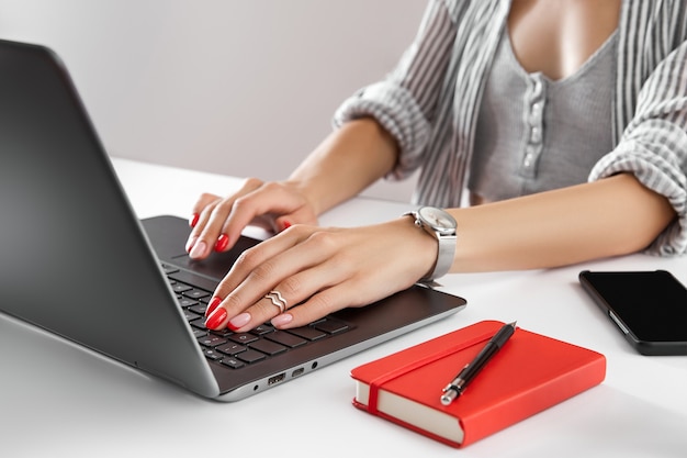 赤いマニキュアを持つ女性は、白いテーブルにノートパソコンを使って仕事をします。