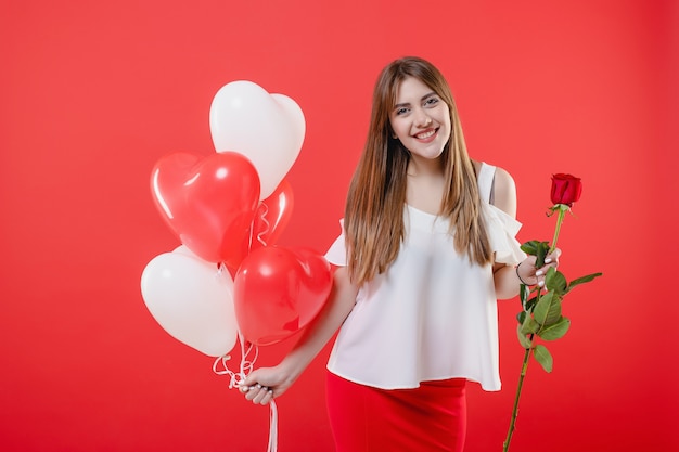 Woman with rose and colorful heart shaped balloons isolated over red wall