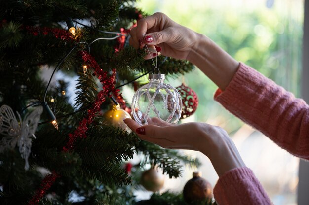 Woman with seasonal manicure hanging shiny transparent holiday bauble on christmas tree.