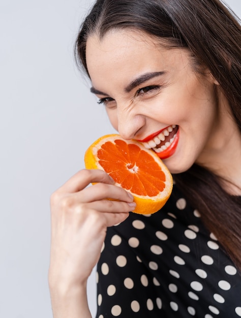 Woman with sensire smile holding half of a grapefruit near red lips. People emotions concept.