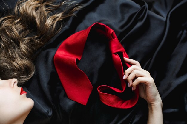 Woman with shiny hair holds a red tie lying on black bed