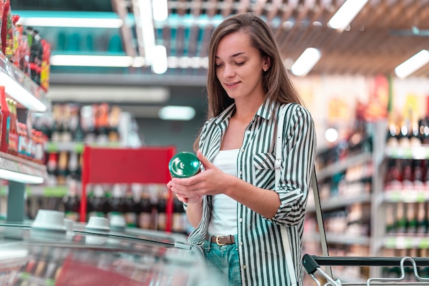 Woman with shopping cart chooses, checking products label and buying food at the grocery shop