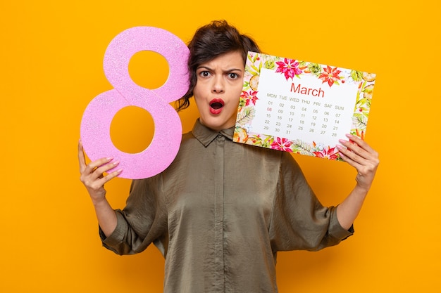 Woman with short hair holding paper calendar of month march and\
number eight looking confused and displeased celebrating\
international women\'s day march 8