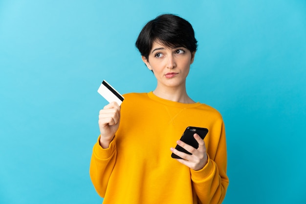 Woman with short hair over isolated buying with the mobile with a credit card while thinking