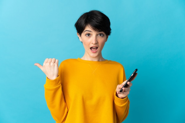 Woman with short hair over isolated wall using mobile phone and pointing to the lateral