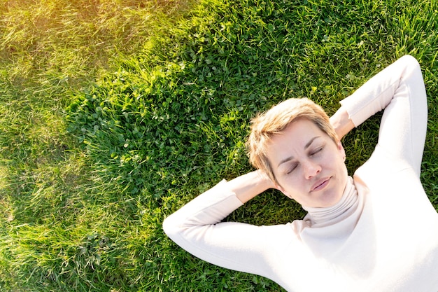 Женщина с короткими волосами в белом топе отдыхает на луговой зеленой траве в солнечный весенний теплый день в парке